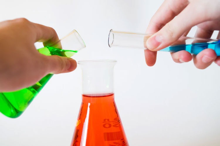 Hands pouring green and blue liquids into a beaker containing a red liquid.